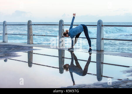 Jeune femme faisant un virage large aux jambes, exercice de corps tordu le long de la plage avec réflexion sur la route mouillée. femme de fitness faisant l'entraînement d'étirement sur humide Banque D'Images