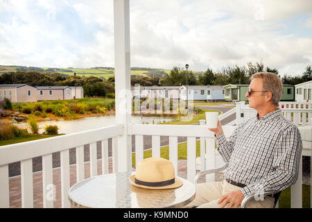 Homme âgé de boire une grande tasse de café assis sur la véranda d'un mobil-home de luxe dans le Nord du Pays de Galles en profitant de la vue panoramique sur le Pays de Galles Banque D'Images