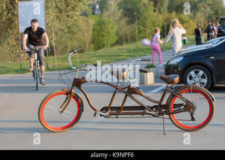 Un tandem couleur chocolat avec des roues est garé dans un parking près de la piste cyclable dans le soleil du soir. Banque D'Images