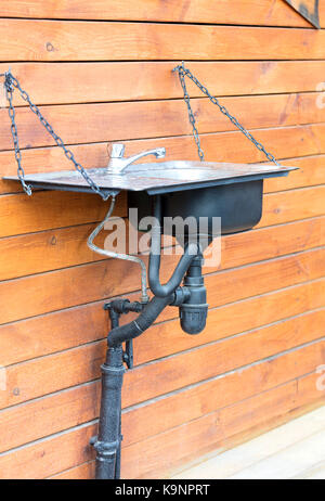 Un lavabo lavabo avec un robinet en acier inoxydable installée à l'extérieur sur une chaîne sur le mur d'une maison en bois, dans une ferme rustique Banque D'Images