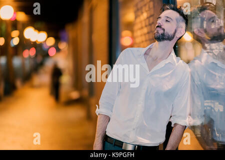 Beau caucasian male professional photographié dans la rue la nuit, appuyé contre une vitrine à la recherche vers le bas. Banque D'Images