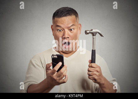 Choqué man looking at smart phone holding hammer isolé sur fond de mur gris Banque D'Images