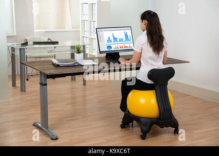 Young businesswoman sitting on fitness ball tout en travaillant sur computer in office Banque D'Images