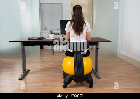 Young businesswoman sitting on fitness ball tout en travaillant sur computer in office Banque D'Images