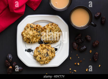 L'avoine et fruits date cookies dans la plaque blanche élégante avec deux tasses de thé sur la table noire avec serviette rouge - Vue de dessus l'arrière-plan de l'alimentation Banque D'Images