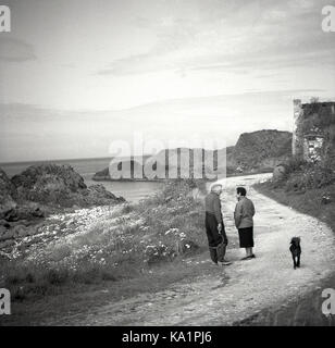 Années 50, 'Toujours demander un local'..Photo historique montrant un homme et une femme ayant une conversation sur un sentier côtier en Irlande du Nord. L'homme, peut-être un agriculteur local, porte des bottes, un chapeau et muni d'un crochet ou d'herbe à la faux à la main. Banque D'Images