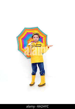 Petit garçon vérifie la présence de pluie alors que vêtue d'un polissoir de jaune et des bottes en caoutchouc et chapeau. il tient un parapluie à rayures dans une chambre toute blanche. Banque D'Images