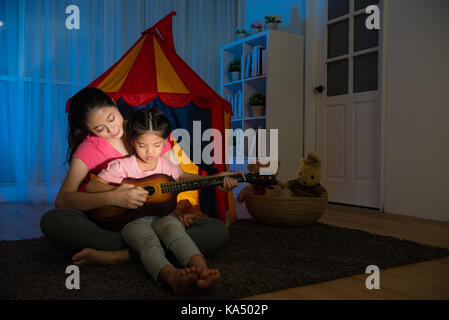 Jeune femme au foyer calme tranquille enseignement belle fille jouer ukulele petite guitare assis sur plancher du salon avec une tente enfants jouets Banque D'Images