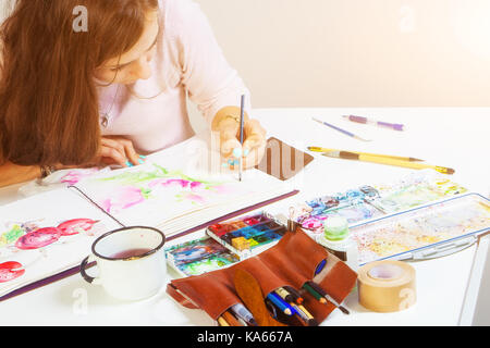 Artiste brune avec des tirages dans un album pour un mince pinceau en bois rose avec des aquarelles, sur la table se trouve un étui en cuir avec des pinceaux, Banque D'Images