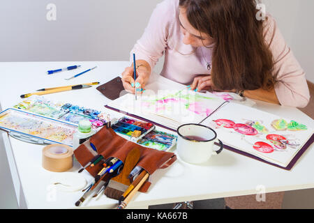 Artiste brune avec des tirages dans un album pour un mince pinceau en bois rose avec des aquarelles, sur la table se trouve un étui en cuir avec des pinceaux, Banque D'Images