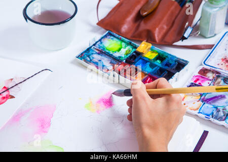 Un close-up d'artiste dessine des fleurs roses avec une brosse en bois et d'aquarelles dans une feuille de travail, sur la table se trouvent un étui en cuir avec des brosses, un drawi Banque D'Images