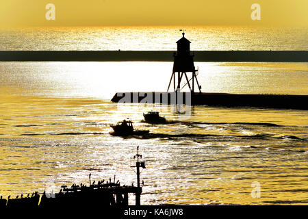 Cormorans sur l'ancienne jetée et les pêcheurs dans leurs bateaux à moteur passant le troupeau phare épi après un lever du soleil d'or Banque D'Images