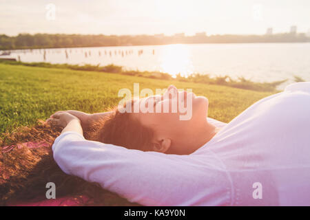 Belle jeune femme enceinte faisant du yoga sur les bords de la rivière. femme allongée sur le dos sur un tapis de yoga. visage féminin, fermé les yeux de près. rivière, Soleil couchant Banque D'Images