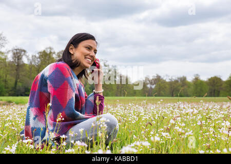 Colombian woman sitting in flowering meadow téléphoner avec un téléphone mobile Banque D'Images