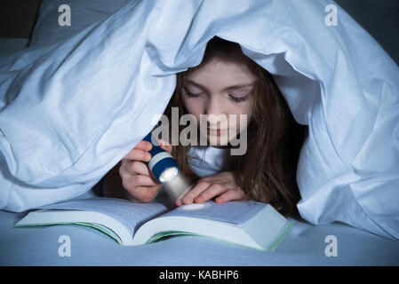 Girl reading book avec lampe de poche sous la couverture sur le lit Banque D'Images