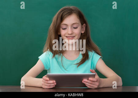 Happy girl holding digital tablet sitting at desk contre green chalkboard Banque D'Images