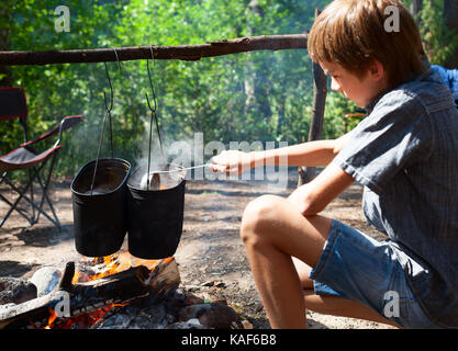 Jeune garçon camp cuisine nourriture dans chaudron sur feu ouvert Banque D'Images