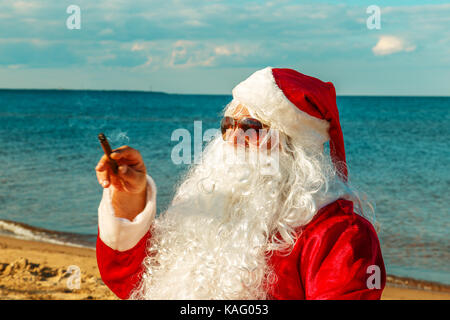Père Noël sur la plage est assis sur une chaise et fume un cigare. Banque D'Images