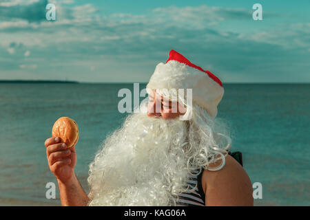 Père Noël sur la plage de manger un hamburger. Le concept de l'alimentation malsaine. Banque D'Images