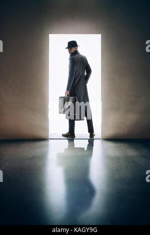 L'homme silhouette en imperméable et chapeau debout dans la lumière de l'ouverture de la porte dans une pièce sombre Banque D'Images