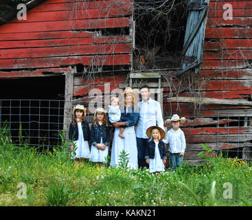 Père et mère posent avec leurs cinq enfants en face d'une grange en bois rustique, rouge. les jeunes filles portent des vestes en denim et les garçons sont vêtus de blanc shi Banque D'Images