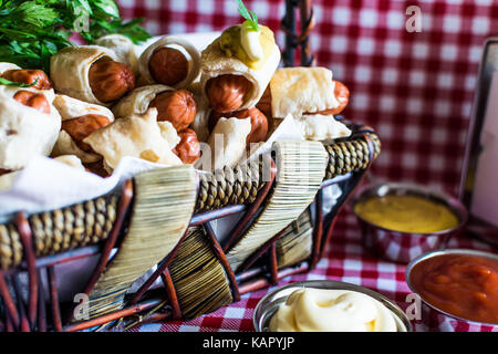 Panier en osier avec mini hot dog (artisan de la saucisse dans la pâte) avec de la moutarde, de la mayonnaise sur un fond à carreaux Banque D'Images