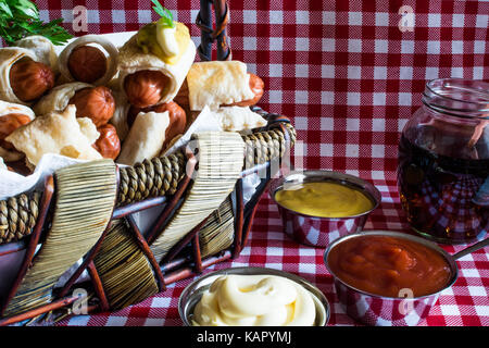 Panier en osier avec artisan mini hot dog (la saucisse dans la pâte) avec de la moutarde, la mayonnaise et le cola sur un fond à carreaux Banque D'Images