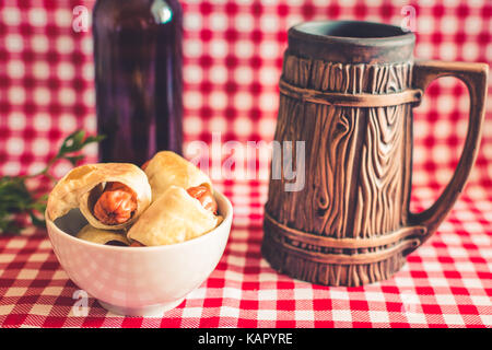 Mini plaque de maison hot dog (saucisse en croûte) sur serviette avec une bouteille de bière brune et faïence mug sur un fond à carreaux Banque D'Images