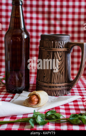 Des mini-saucisses hot-dog (en pâtisserie) sur la serviette avec une bouteille de bière brune et faïence mug sur un fond à carreaux Banque D'Images