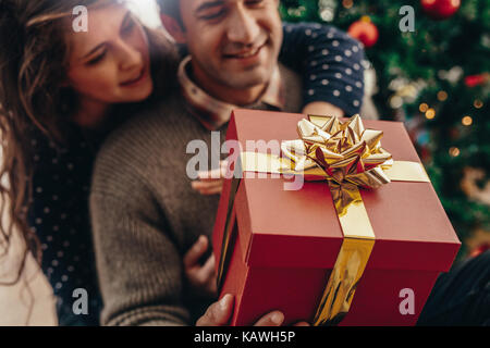 Jeune homme et femme avec une boîte cadeau joliment décorées, assis près de l'arbre de Noël. Heureux couple célébrer Noël avec des cadeaux. Banque D'Images