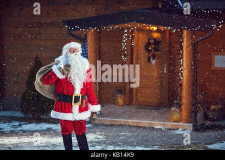 Père Noël avec sac plein de cadeaux dans l'avant de la maison en bois Banque D'Images