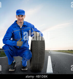 Young smiling mechanic showing Thumbs up next les pneus de voiture - service de remorquage, l'assistance routière Banque D'Images