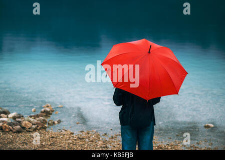 Femme avec parapluie rouge prévoit de la pluie en face d'un lac. triste et solitaire à la personne de sexe féminin dans la distance. Banque D'Images