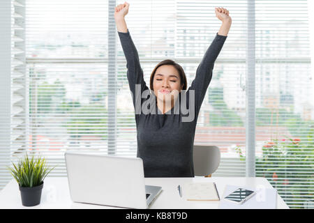 Bonne ambiance jeune femme assise à son bureau avec un ordinateur portable, s'étirant ses bras Banque D'Images