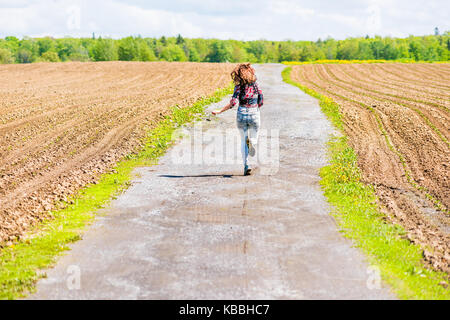 Jeune femme courir, sauter dans l'air et souriant sur la route de campagne par brown champs labourés avec sillons en été à l'île d'Orléans, Québec, Canada Banque D'Images