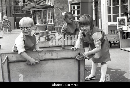 70, tableau historique de trois professionnels de jeunes enfants portant des bretelles à l'aide de brosses pour nettoyer une table à l'extérieur dans une pépinière sur un ensemble immobilier conseil à Deptford, le sud de Londres, en Angleterre. Banque D'Images