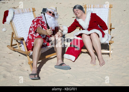 Le Père Noël en rouge les slips et Hawaiian shirt se prélasser sur une plage de sable avec Mrs Claus prendre. selfies Banque D'Images