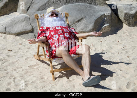 Père Noël en maillot de bain rouge et chemise hawaïenne se prélasser sur une plage de sable avec chapeau de paille et lunettes de soleil Banque D'Images