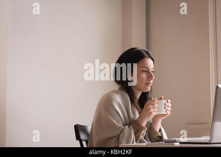 Jeune femme à la recherche d'ordinateur portable en tout en ayant le café à la maison Banque D'Images