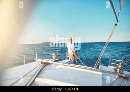 Smiling mature man seul se tenant sur le pont de son bateau avec la gouverne de direction tout en naviguant sur l'océan ouvert sur une journée ensoleillée Banque D'Images