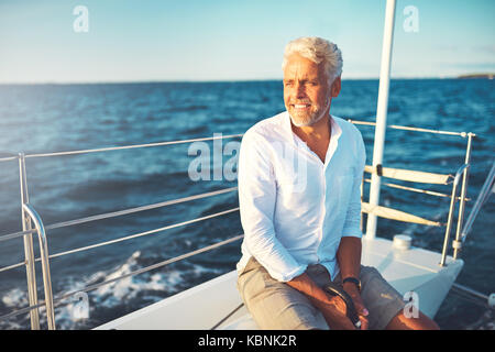 Smiling mature man assis tout seul sur le pont de son bateau bénéficiant d'une journée ensoleillée de la voile sur l'océan Banque D'Images