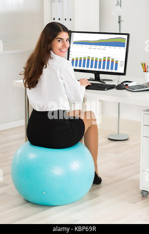 Portrait of happy young businesswoman using computer while sitting on exercise ball in office Banque D'Images