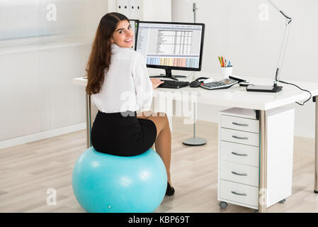 Portrait of happy young businesswoman using computer while sitting on exercise ball in office Banque D'Images