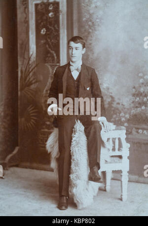 American archive monochrome studio portrait photographie d'un jeune homme portant un costume et un noeud papillon perché sur une chaise, prise à la fin du XIXe siècle, États-Unis Banque D'Images