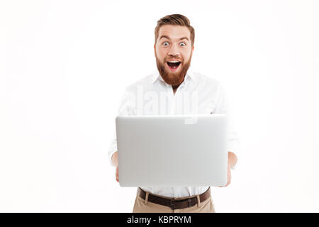 Photo de choqué happy young bearded man standing over white background Mur isolé. À la caméra à l'aide d'ordinateur portable. Banque D'Images