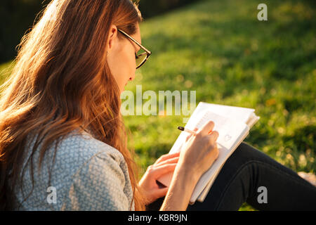 Close up side view image de femme brune à lunettes assis près de l'arbre dans le parc et d'écrire quelque chose sur un ordinateur portable Banque D'Images