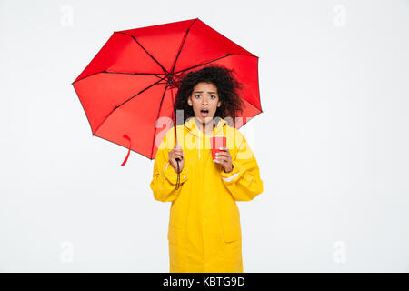 Choqué femme africaine en se cachant sous l'imperméable tout en maintenant parapluie tasse et regardant la caméra Banque D'Images