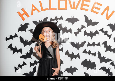 Funny little girl habillé en costume halloween citrouille holding mask et posant avec les chauves-souris sur un arrière-plan Banque D'Images