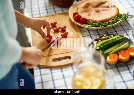 Gros plan du jeune femme douce coupe tomates cerise et d'autres légumes sur planche de bois tandis que la préparation d'un repas avec des amis à la table rustique Banque D'Images