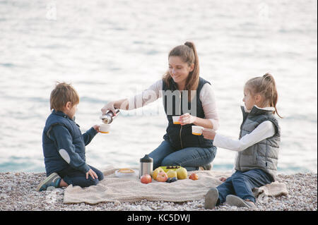 Smiling belle mère remplissant les enfants tasses avec du thé chaud assis au bord de la mer au début de l'hiver Banque D'Images